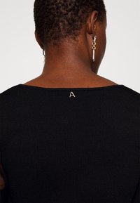 Schwarzes, geripptes Oberteil mit weitem Ausschnitt, das auf der Rückseite mit einem kleinen, silbernen Buchstaben "A" bestickt ist. Die Haare sind kurz und eng geschnitten.