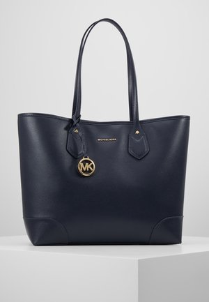 Granatowa skórzana torba typu tote Michael Kors z podwójnymi paskami na ramię i złotym zawieszką z logo MK wiszącą w pobliżu górnego środka.