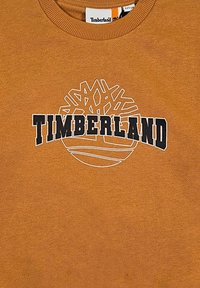 Oransje genser med rund hals som har et hvitt logo av et tredesign over teksten "TIMBERLAND" i svart. Stoff i bomullsblanding.