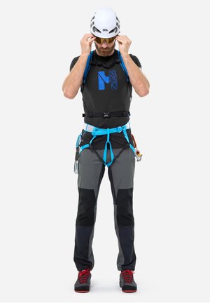 Homme ajustant ses lunettes de soleil, portant un casque d'escalade blanc, un harnais d'escalade noir et bleu, un pantalon noir et gris, et des chaussures d'escalade à lacets rouges.