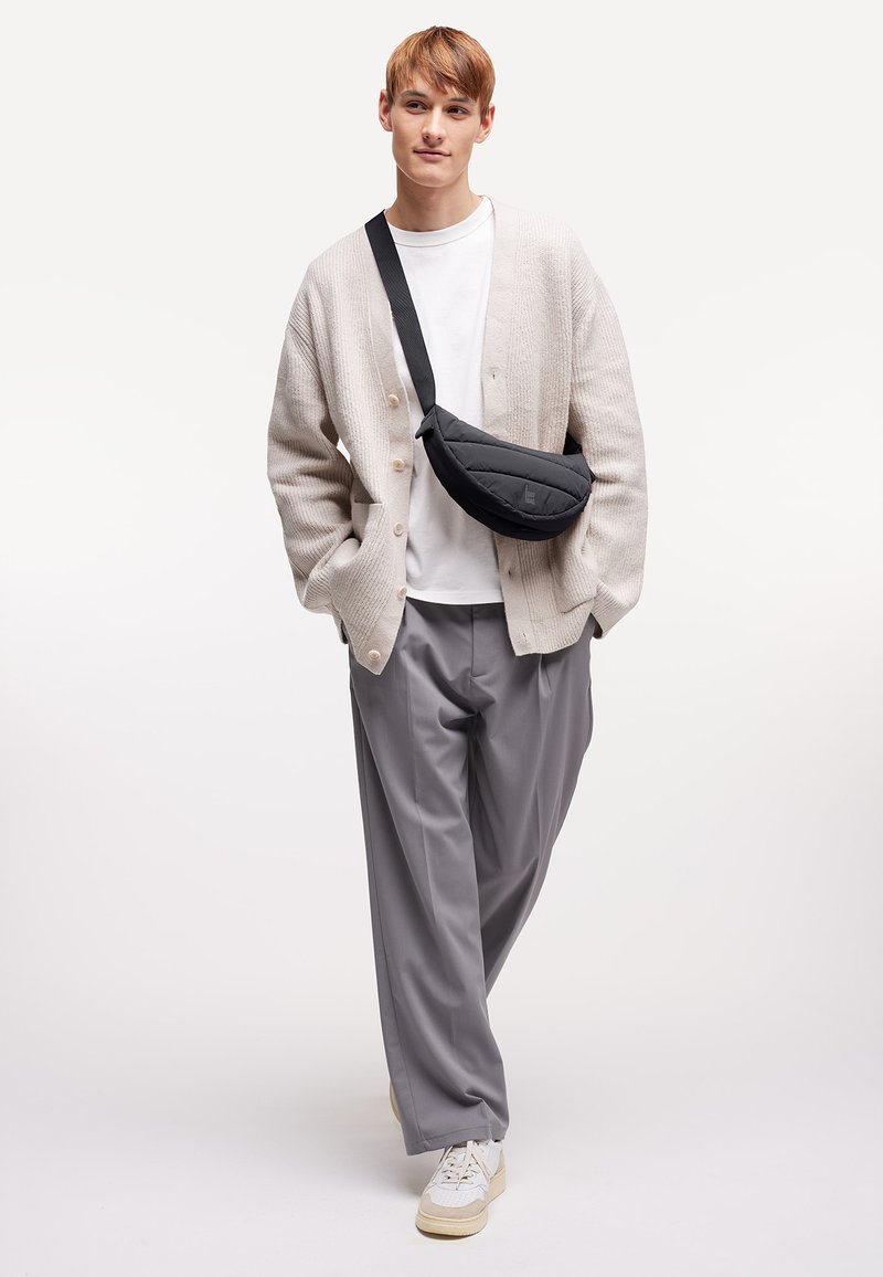 Ung mand med lysebrunt hår iført beige cardigan, hvid T-shirt, grå vide bukser, hvide sneakers og sort skuldertaske står på hvid baggrund.