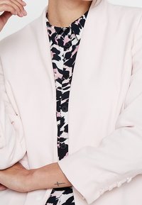 Světlé růžové sako s šálovým límcem, dlouhými rukávy a knoflíkovými detaily. Nosené přes černou košili s růžovým vzorem a knoflíky.
