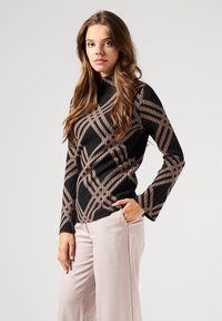 Maglia a maniche lunghe a righe nere e marroni con un motivo geometrico, colletto alto e silhouette aderente, abbinata a pantaloni di colore chiaro.