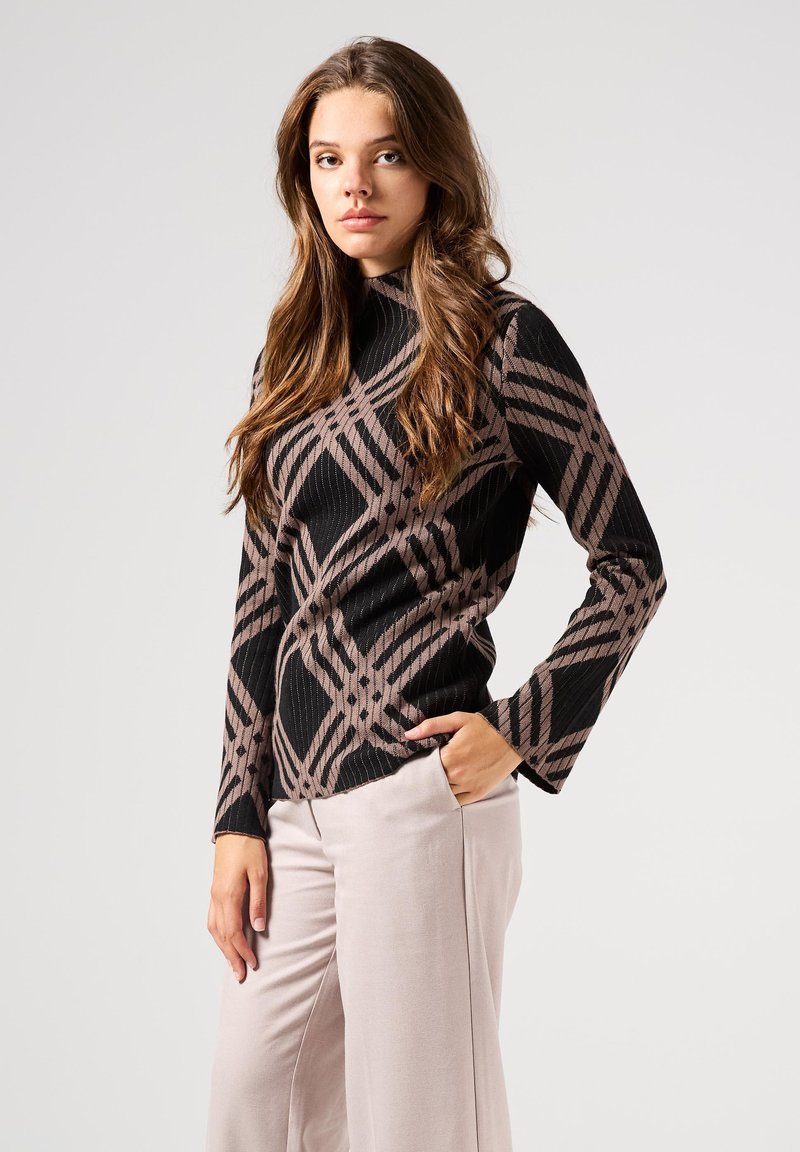 Maglia a maniche lunghe a righe nere e marroni con un motivo geometrico, colletto alto e silhouette aderente, abbinata a pantaloni di colore chiaro.