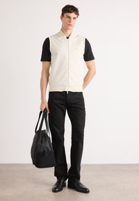 Gaišas bezpiedurkņu veste virs melnas T-kreklu, apvienota ar melnām biksēm un pulētiem apaviem. Modelis tur melnu somu, stāvot uz neitrāla fona.