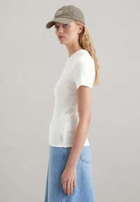 Weißes, geripptes Kurzarmoberteil, kombiniert mit einem hellblauen Jeansrock. Das Modell trägt eine olivgrüne Mütze mit weißem Text.