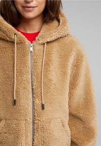 Femme portant un sweat à capuche zippé beige et duveteux avec des cordons, par-dessus un t-shirt rouge, visible des épaules à la poitrine sur un fond gris.