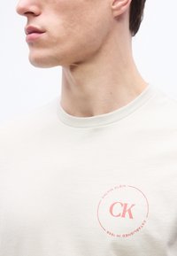 Κοντινό πλάνο ενός ατόμου που φοράει λευκό t-shirt με ροζ λογότυπο «CK Calvin Klein Established in 1968» στο στήθος.