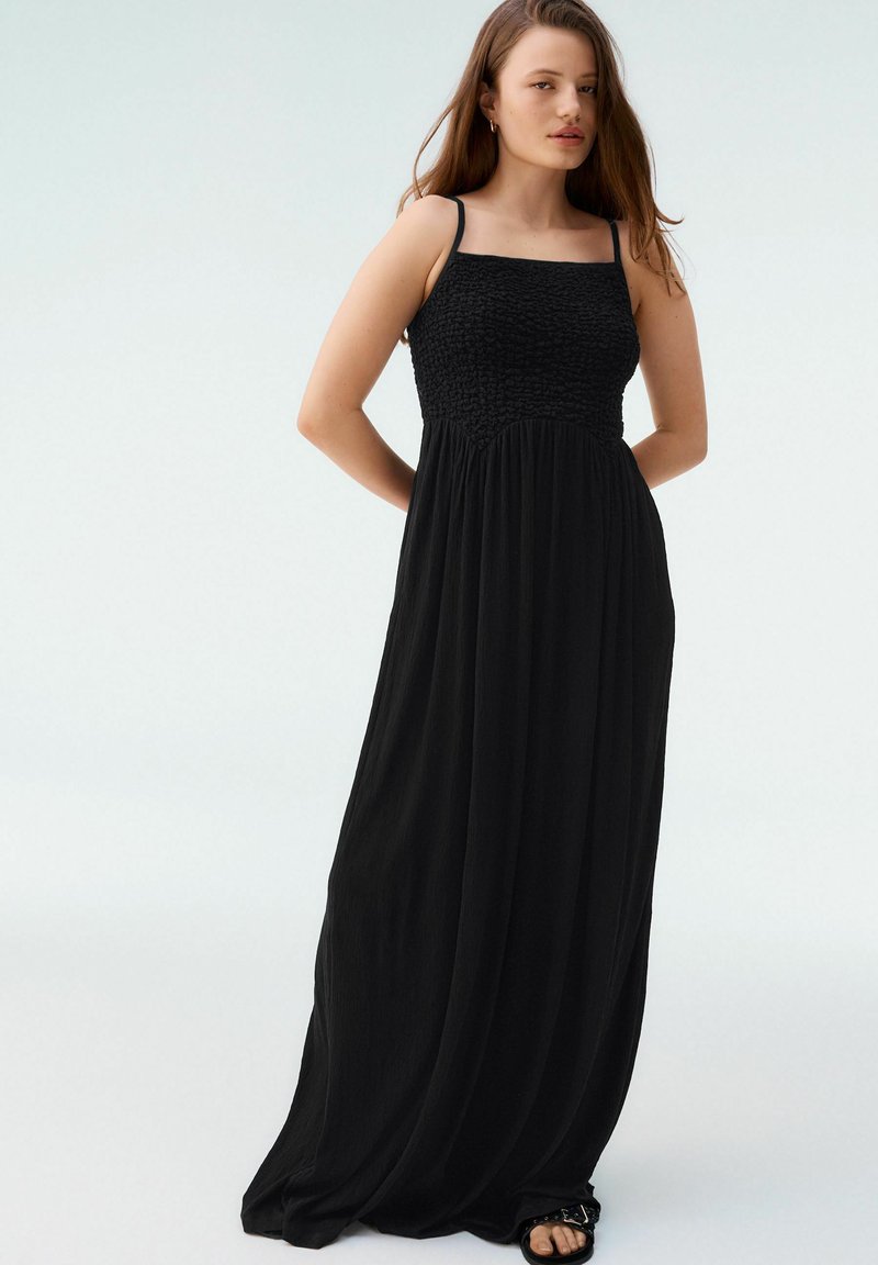 Robe noire maxi avec un corsage smocké, décolleté carré, fines bretelles et jupe fluide. Tissu léger avec une texture douce.