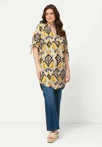Lange, beige tuniek met veelkleurige geometrische patronen, met gestrikte mouwen en een v-hals, gecombineerd met een wijde denim broek en platform sandalen.