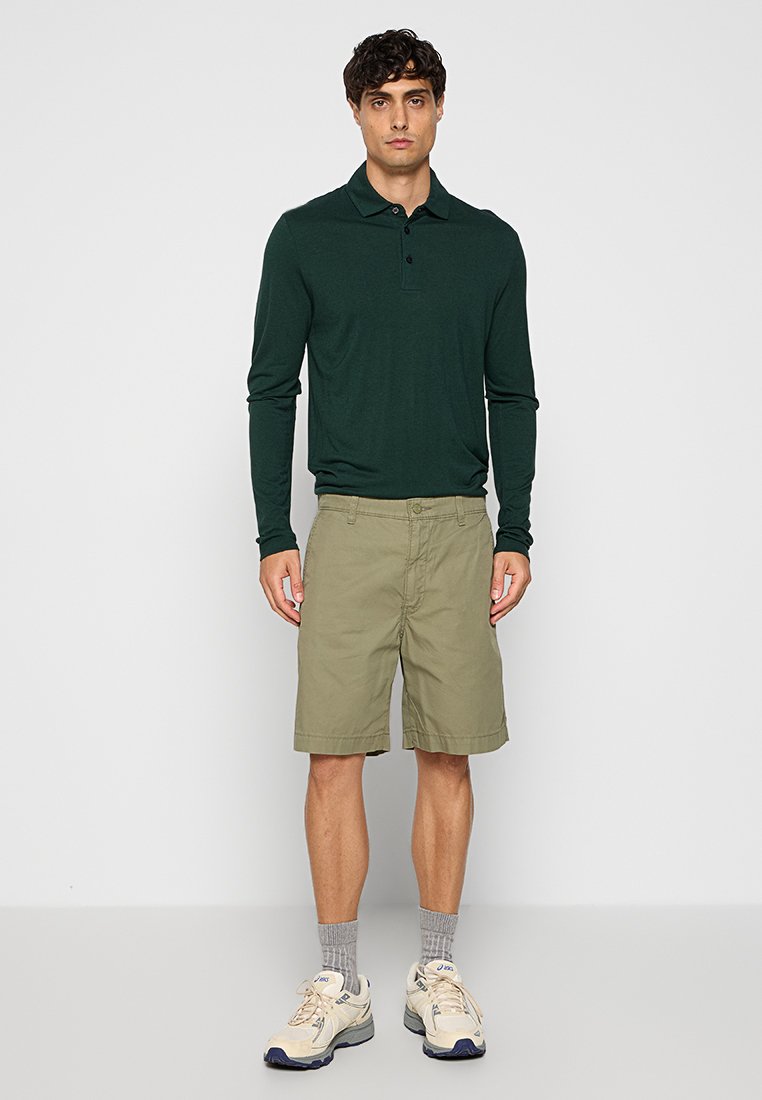 Langärmliges dunkelgrünes Polohemd, kombiniert mit khakifarbenen Shorts. Das Outfit umfasst beigefarbene Sneakers und graue Socken, die ein schlichtes Design aufweisen.