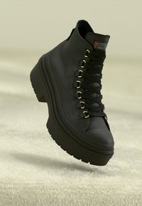 Bota negra con cordones, suela gruesa, ojales metálicos y textura de cuero. Presenta una punta redondeada y altura de tobillo corta.