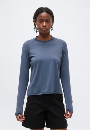 Mladá osoba s vrkočmi má na sebe dlhý modrý rukáv a čierne šortky, stojí pred jednoduchým svetlým pozadím.