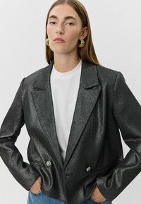 Zwart metalen blazer met een gestructureerde afwerking, met een notch-kraag, dubbelgeknoopte ontwerp en knopen met bloemdetails; gecombineerd met een wit shirt.