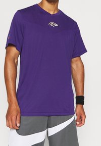 Fialové sportovní tričko s krátkými rukávy a výstřihem do V, s logem Baltimore Ravens na hrudi. Hladká tkanina v jednotné barvě.