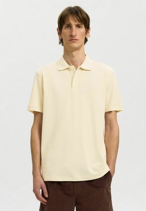 Homme aux cheveux courts et bruns portant un polo jaune pâle à manches courtes et un pantalon marron, debout devant un fond uni.