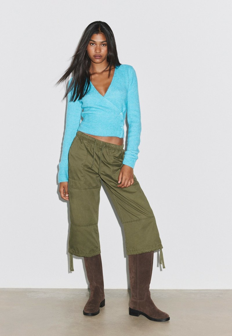 Jeune femme aux longs cheveux foncés porte un pull court bleu, un pantalon large olive et des bottes marron montant jusqu'aux genoux, debout contre un mur blanc.