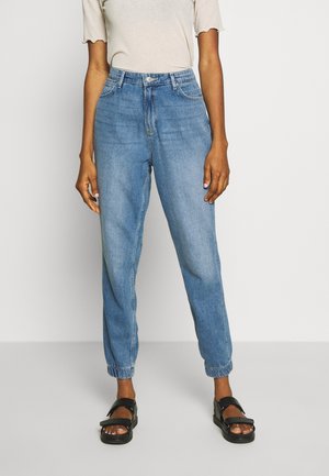 Person trägt hellblaue Denim-Jogginghosen mit elastischen Bündchen und ein beiges kurzärmeliges Oberteil und steht vor einem einfarbigen Hintergrund.