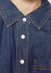 Denimová košile s špičatým límcem, ozdobená perlami jako knoflíky, oranžovým prošíváním a náprsní kapsou. Textura je hladká s klasickým modrým odstínem.