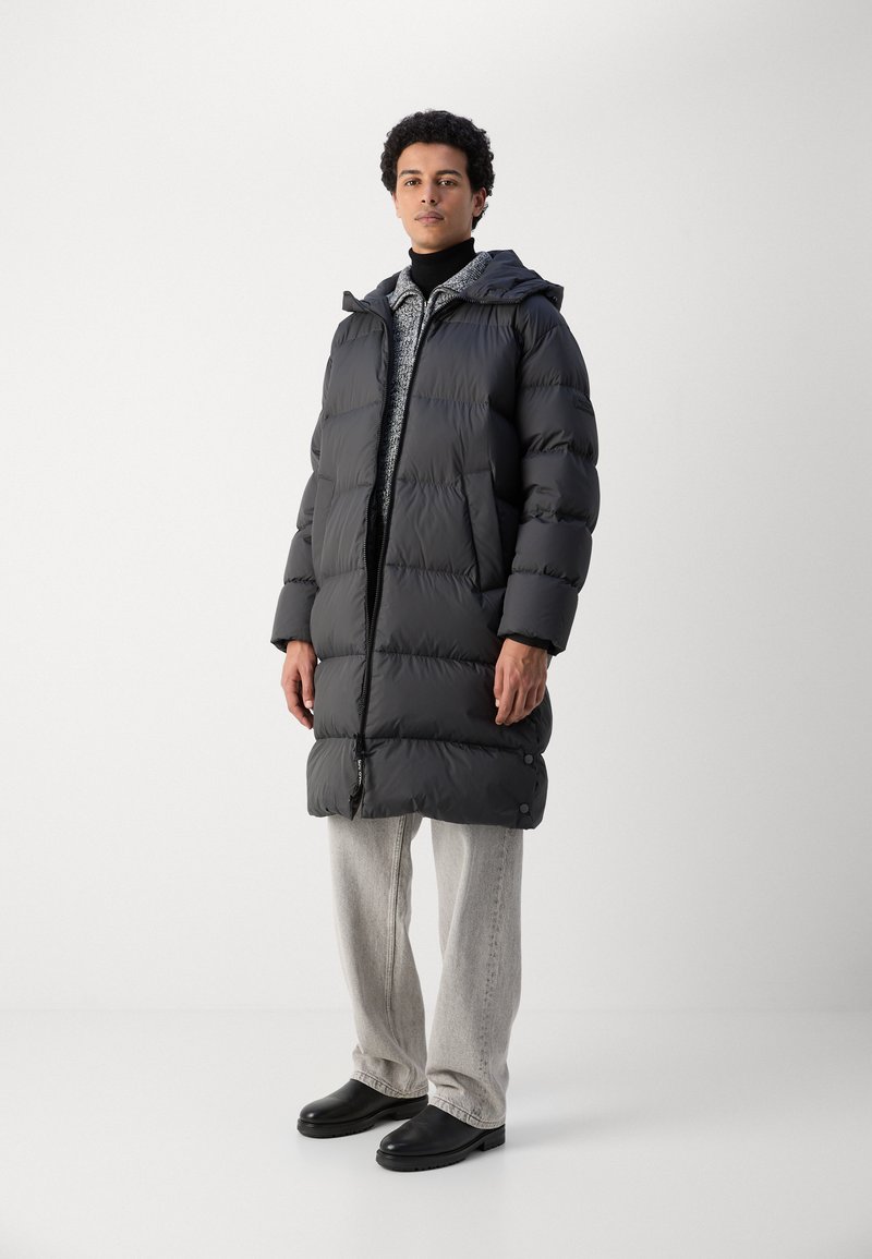 Abrigo tipo puffer negro con capucha, diseño acolchado, cierre de cremallera y botones a presión. Llevado sobre un suéter ligero texturizado y pantalones gris claro.