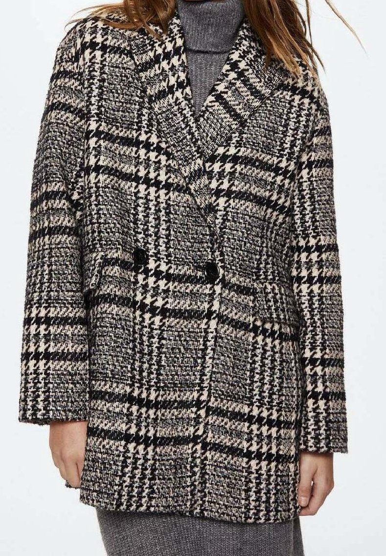 Femme portant un manteau croisé à motif pied-de-poule noir et blanc sur une robe en tricot gris à col roulé.