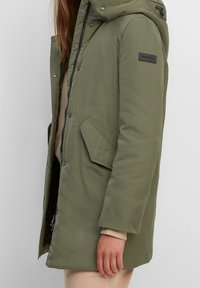 Parka verde oliva con cappuccio, chiusura frontale con zip e bottoni a pressione, tasche laterali e tessuto testurizzato. Presenta una toppa con il marchio sulla manica.