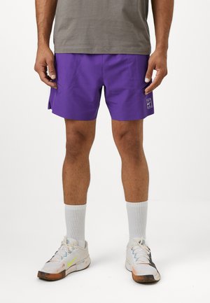 Lila Sportshorts mit Seitenöffnungen, aus leichtem Material. Getragen mit weißen Socken und Sportschuhen mit bunten Akzenten.