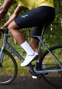 Vélo de canyon gris avec des composants noirs. Le cycliste porte un maillot jaune, un short noir avec le logo "Rapha" en blanc, des chaussettes blanches et des chaussures de cyclisme blanches.