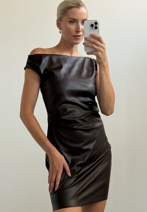 Femme aux cheveux clairs tirés en arrière, portant une robe noire courte, brillante et sans épaules, tenant un smartphone blanc en train de prendre un selfie dans le miroir.