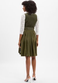 Frau mit lockigem Haar, die ein olivgrünes, plissiertes Kleid mit weißen Spitzenärmeln und silbernen High-Heels trägt und seitlich schaut.