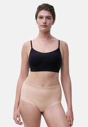 Frau trägt einen schwarzen nahtlosen Bralette und hautfarbene High-Waist-Unterwäsche mit Spitzenabschluss, steht vor einem schlichten hellen Hintergrund.