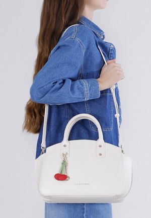 Mujer con una chaqueta de mezclilla azul lleva un bolso blanco con doble asa y un colgante en forma de cereza sobre su hombro.