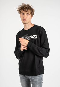 Sudadera negra con cuello redondo y puños de canalé, con letras blancas en la parte frontal. El modelo lleva puestos jeans grises desgastados.
