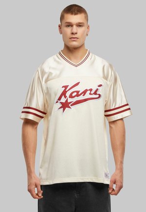 Jersey de couleur crème avec une texture en maille, arborant un logo imprimé rouge et un motif d'étoile. Accents rayés rouges sur les manches. Forme en V.
