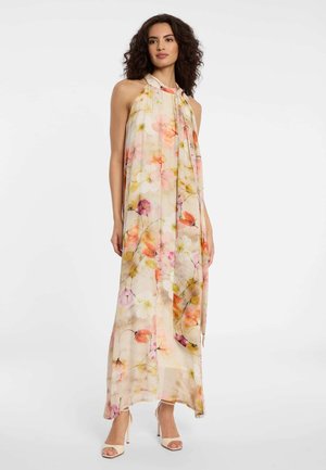 Frau trägt ein ärmelloses, neckholder-floral Maxikleid mit hellrosa und orangefarbenen Blumenmustern, kombiniert mit weißen Sandaletten mit hohen Absätzen.