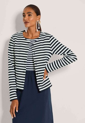 Frau mit dunklen Haaren im Pferdeschwanz trägt eine schwarz-weiß horizontal gestreifte, geknöpfte Jacke über passendem Oberteil und dunklem Rock und posiert mit der Hand in der Hüfte.