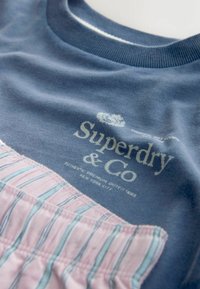 Modrá bavlněná tričko s texturovanou růžovou kapsou se proužky, s bílým nápisem "Superdry & Co" vytištěným na hrudi.