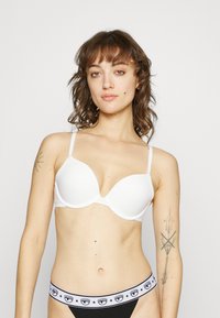Sujetador blanco con copas lisas, tirantes finos ajustables y un diseño sin costuras. Combinado con ropa interior negra que tiene una cinturilla con patrón.