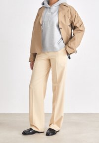 Trench beige sopra un hoodie grigio, abbinato a pantaloni larghi beige e mocassini neri. Tessuto morbido e vestibilità rilassata in tutto.