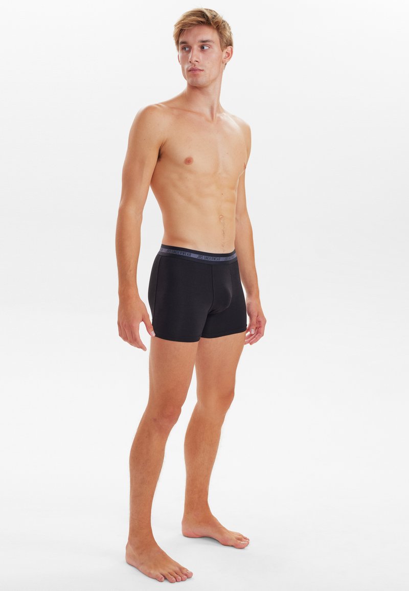 Sorte boxer-shorts i blødt stof, tætsiddende design og elastisk talje med subtil branding. Modellen poser neutralt.
