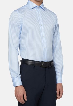 Mann trägt ein hellblaues, vertikal gestreiftes Hemd, das in dunkelblaue Hose mit schwarzem Ledergürtel gesteckt ist, und steht vor einem neutralen Hintergrund.