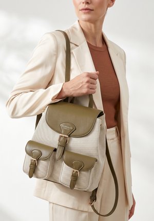 Donna in completo beige che tiene uno zaino beige e verde oliva Diana & Co Firenze con due tasche anteriori con fibbie.