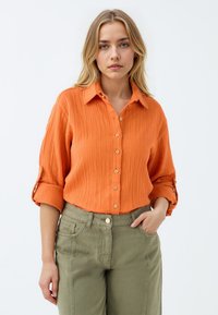 Oranges strukturiertes Button-up-Hemd mit hochgekrempelten Ärmeln, kombiniert mit olivgrünen Hosen, die über Fronttaschen und ein Metallknopfdetail verfügen.