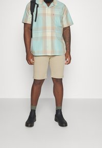 Camicia a maniche corte a scacchi blu chiaro e beige, pantaloni corti beige, stivali neri alla caviglia, calzini verdi e uno zaino nero. Colorazioni uniche e motivi.