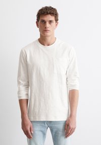 Weißes Langarm-T-Shirt aus weichem Stoff, mit rundem Halsausschnitt und einer linken Brusttasche. Trägt man mit hellblauen Jeans.