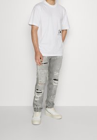 Mies, joka on pukeutunut valkoiseen Calvin Klein -t-paitaan, vaaleanharmaisiin revittyihin farkkuihin mustilla paikoilla ja valkoisiin lenkkareihin, seisoo valkoista taustaa vasten.