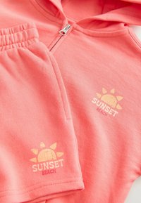 Korallowy różowy zestaw z kapturem i szortami wykonany z miękkiej tkaniny, z zamkiem błyskawicznym oraz grafiką słońca z napisem "SUNSET BEACH" w jasnym żółtym kolorze.