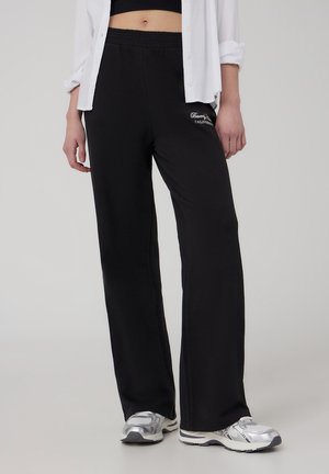 Pantaloni sportivi neri con elastico in vita, dotati di un dettaglio con logo. Abbinati a una camicia bianca con bottoni e scarpe da ginnastica argentate.