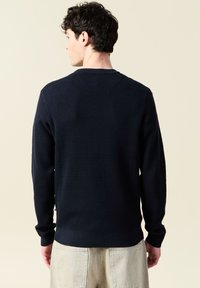 Navyblå texturerad tröja, rund hals, ribbade mudd och nederkant, med en åtsittande silhuett och fint stickat mönster, bärs över ljusa byxor.