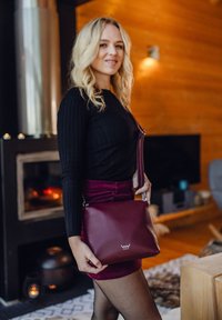 Bordová kožená kabelka přes rameno s hladkým povrchem a logem. Kabelka má dlouhý popruh a elegantní obdélníkový tvar.